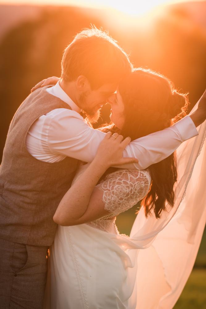 Heterosexuelles Hochzeitspaar Nase an Nase im Sonnenuntergang. Er beugt sich zu ihr und hält ihre Schleppe hoch.